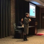 A woman with a microphone is speaking, while another woman is seated at a table behind her and listening intently